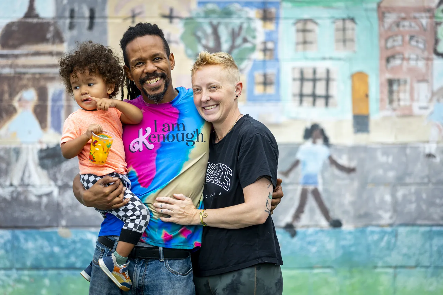 A dad holding his toddler, smiling with his partner in front of a painted mural