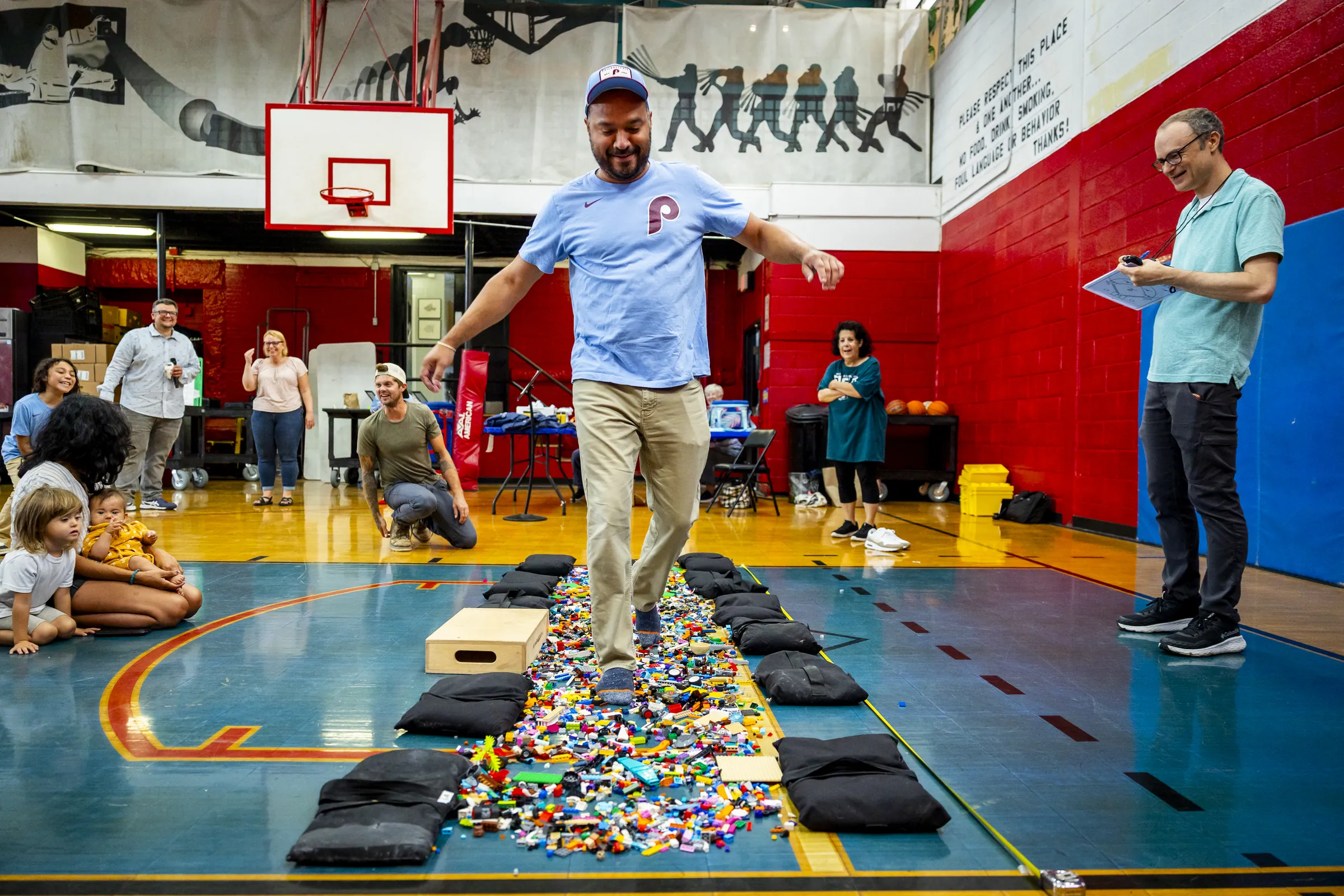 A dad walks barefoot down a path of LEGO bricks in a school gym while families watch